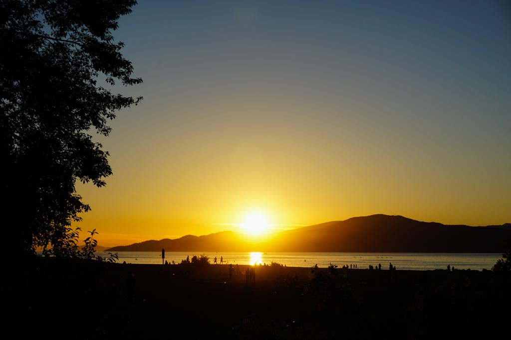 A orange sunset at Spanish Banks beach in Vancouver. The sun is setting on the mountains and reflecting on the water next to the beach.